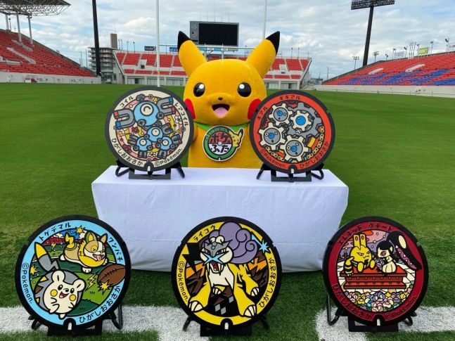 Imagem: Pikachu em estádio de Osaka, atrás de mesa com dois modelos de bueiro (Magnemite + Magnetom, e Kinkl + Klang), e no chão mais 3 modelos (Togedemaru + Yamper, Elekid + Mawile, Raikou).
