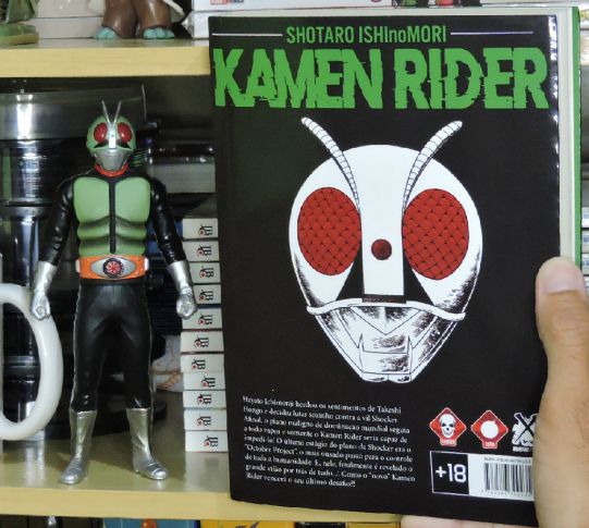 Imagem: Capa traseira do volume 3 de 'Kamen Rider' ao lado de boneco do personagem.