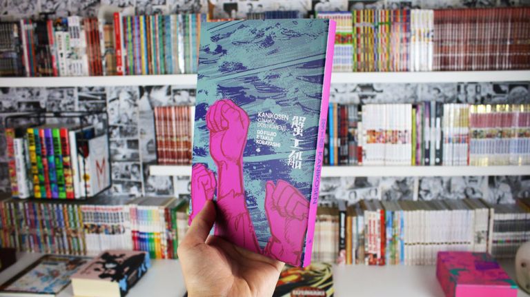 Imagem: Foto de mão segurando capa do Kanikosen, ao fundo uma estante cheia de livros e mangá.