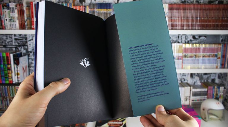 Imagem: Foto de mão abrindo a primeira págima e mostrando a contracapa de Kanikosen, ao fundo uma estante cheia de livros e mangá.