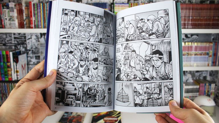 Imagem: Foto de mão segurando o Kanikosen mostrando páginas da história, ao fundo uma estante cheia de livros e mangá.