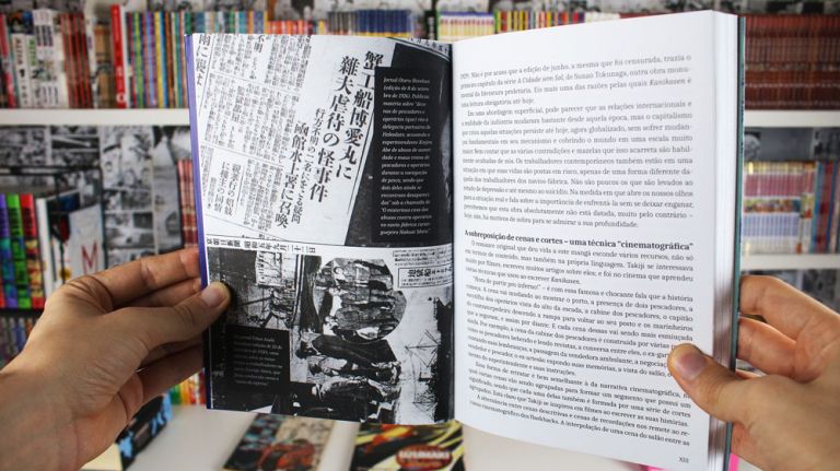 Imagem: Foto de mão segurando o Kanikosen mostrando páginas do prefácio, ao fundo uma estante cheia de livros e mangá.