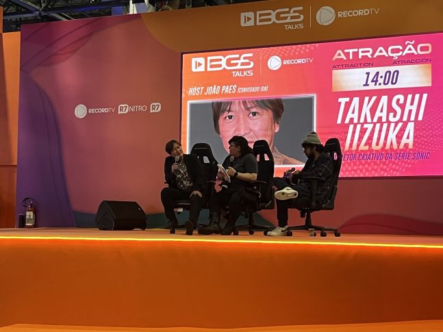 imagem: Takashi Iizuka dando uma entrevista em um palco na Brasil Game Show 2023. Ao lado dele, está a tradutora e o entrevistador